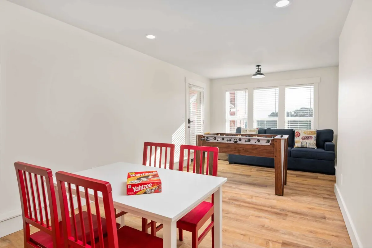 A spacious, cheerful room with a foosball table, red dining chairs around a white table, a blue couch, and large windows letting in daylight. The layout encourages group interaction, meals, and games.