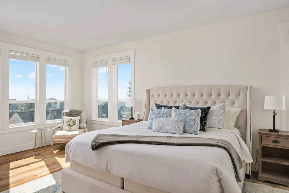 A serene bedroom featuring a plush bed, large windows allowing in natural light, an armchair, and a nightstand. The atmosphere is peaceful and inviting, perfect for morning coffee or a quiet moment of relaxation. The decor is soft and understated, emphasizing comfort and tranquility.