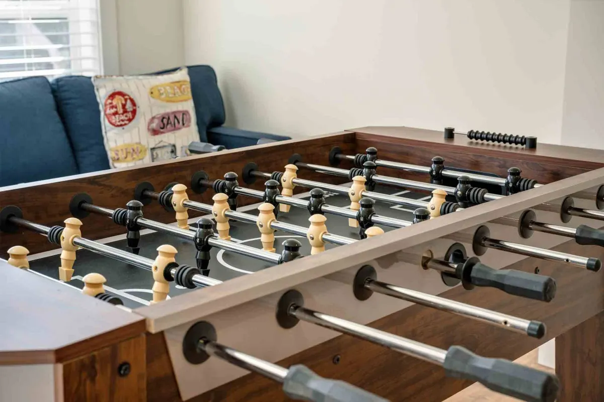 A vibrant game room scene featuring a foosball table, a bright blue couch, and playful decorative pillows. The room radiates energy and is clearly set up for active fun and social interaction.