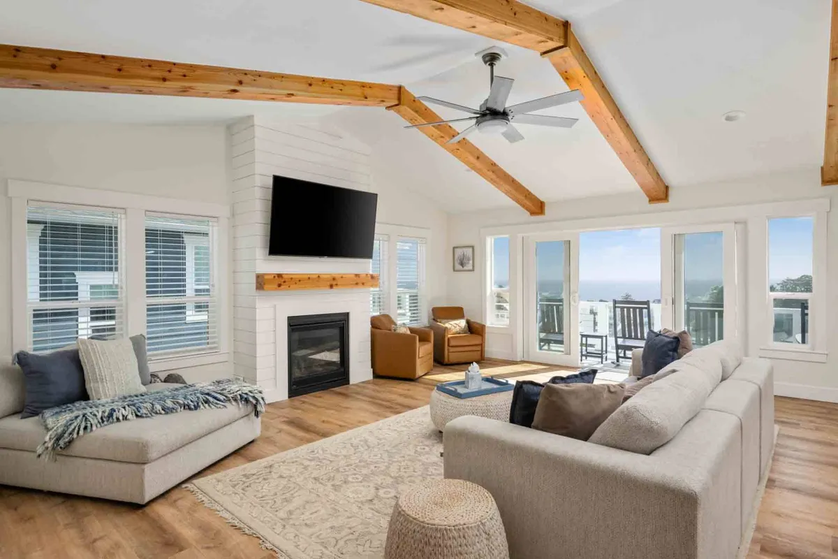 The image showcases a spacious, sunlit living room featuring a large sectional sofa arranged around a cozy fireplace. Massive windows frame stunning ocean views, while exposed wooden beams add warmth and character. The setting is perfect for gathering with family or friends, watching movies on the flat-screen TV, or simply soaking up the coastal ambiance.