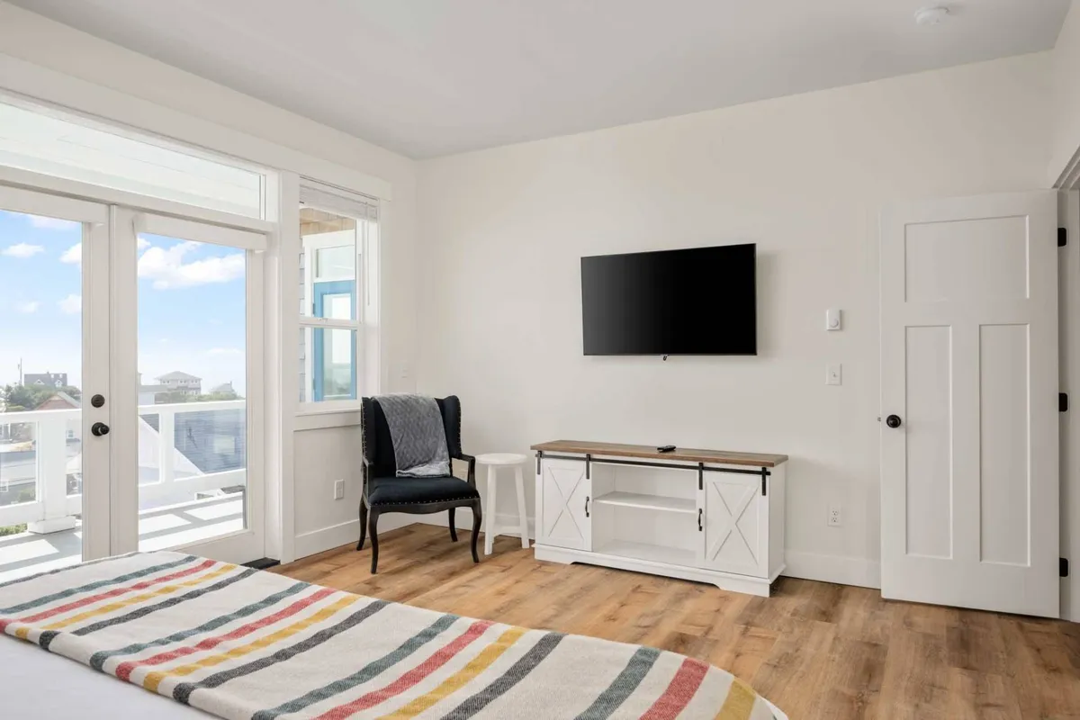 A bedroom featuring a striped bedspread, a TV mounted on the wall, French doors opening to a balcony, and a cozy chair. The room is bright and airy, offering easy access to outdoor views and fresh air.