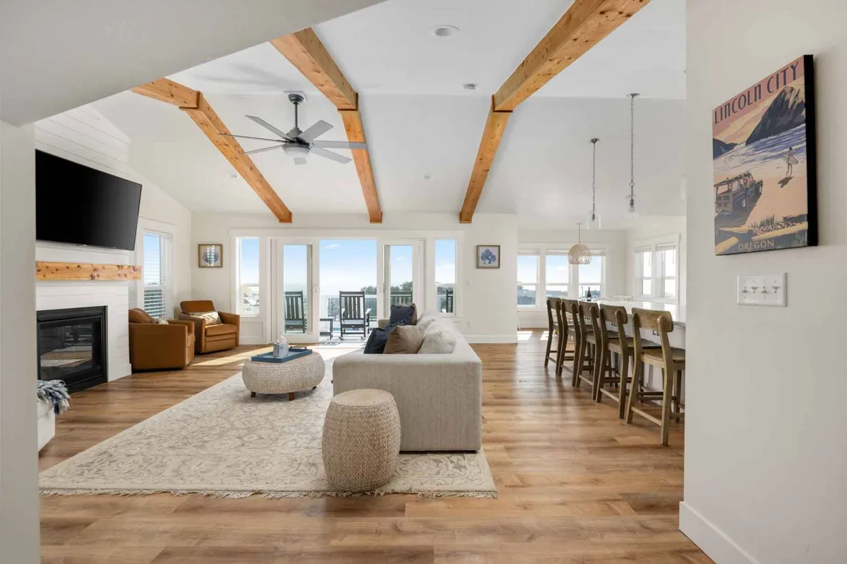 A bright interior with large windows, a comfortable seating area, a stylish dining table, and decorative artwork. The room suggests a warm environment for family gatherings, casual dining, and relaxing by the fireplace.