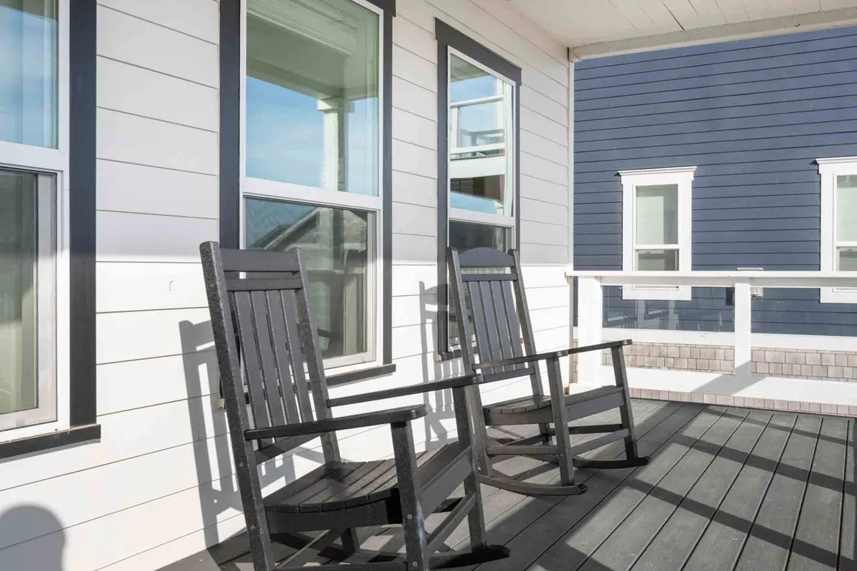 A spacious deck outfitted with rocking chairs, surrounded by railings and large windows, offers a peaceful space to enjoy the breeze and coastal vistas. The calm, serene mood is perfect for unwinding or enjoying a good book.