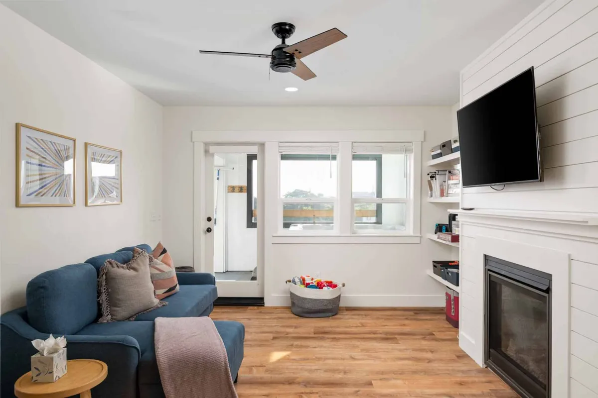 A comfortable living space featuring a blue sofa, a gas fireplace, a large TV, and generously sized windows. The décor is warm and inviting, with ample seating for family and friends to relax or play games together.