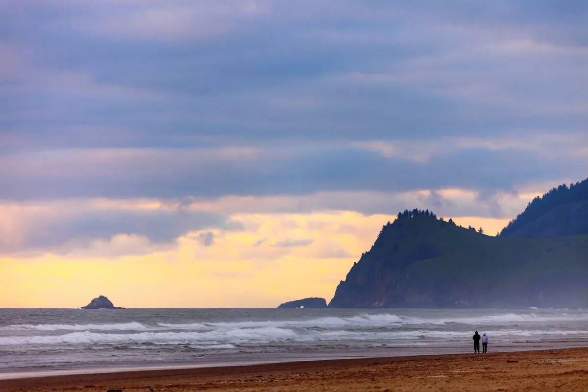 A stunning exterior shot of a couple walking along the sandy beach, with distant cliffs and gentle waves rolling in under a vivid sunset sky. The ocean looks calm and inviting, showcasing the vast natural beauty of the Oregon Coast.