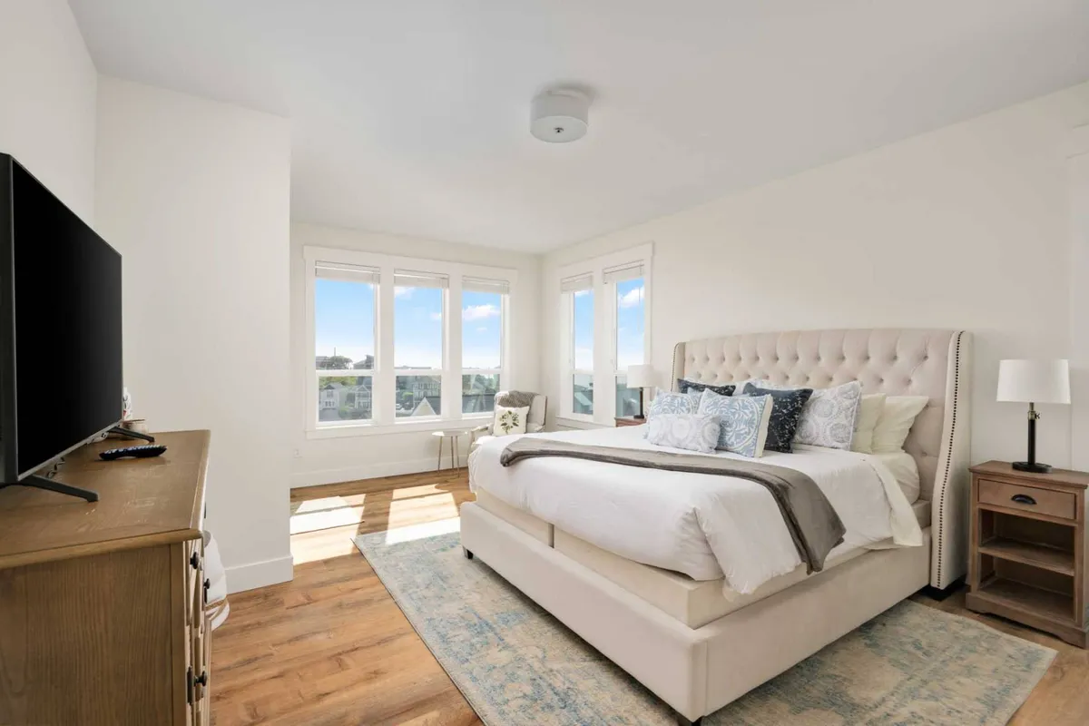 A cozy bedroom with a plush headboard, decorative pillows, a large window filling the room with natural light, and a television atop a dresser. The overall feel is serene and inviting, designed for restful nights and leisurely mornings.